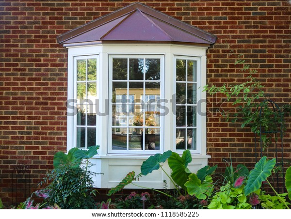 Bay window in a brick house with reflection of\
trees and view of windows and flowers inside and flowers and\
elephant ears outside