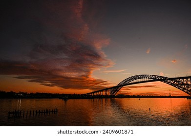 Bayonne Bridge at sunset with dramatic clouds, orange color reflecting on water 