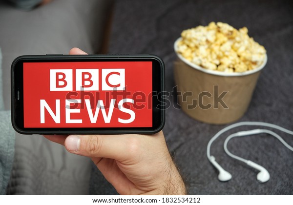 BBC News logo on the smartphone\
in mans hand with popcorn box and apple earphones on the\
background. Advertising concept of news. August 2020, San\
Francisco, USA