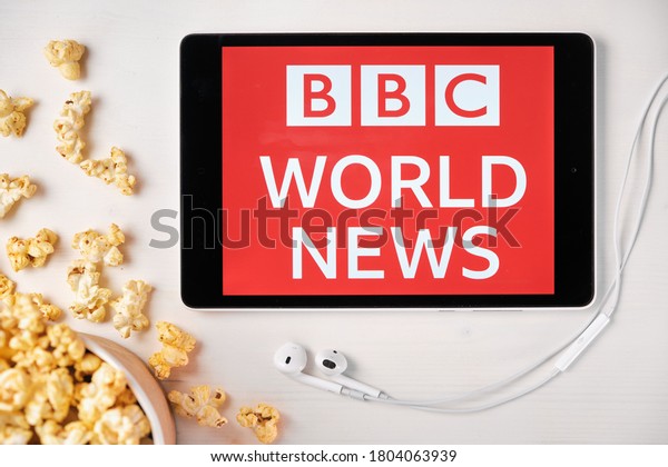 BBC\
World News logo on the screen of the tablet laying on the white\
table and sprinkled popcorn on it. Apple earphones near the tablet\
showing BBC app, August 2020, San Francisco,\
USA