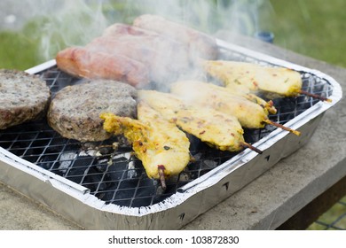 BBQ - Burgers, Satay And Sausages On A Lit Disposable Barbecue.