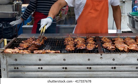 BBQ Chicken Lined Up On A Large Outdoor Grill With Smoke Coming Up From The Fire Below. Person In Orange Apron White Shirt Gloved Hand Holding Tongs Turning Meat