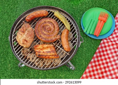 BBQ Portable Grill With Assorted Meat And Picnic Red Blanket On The Backyard Lawn. Overhead View. Summer Outdoor Barbecue Party Or Picnic Concept.