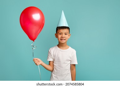 B-Day Celebration. Portrait Of Excited Happy Little Asian Boy Holding Red Baloon, Cheerful Korean Male Kid Wearing Party Hat Celebrating Birthday, Posing Over Blue Studio Background, Copy Space