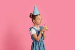 B-Day Wish. Cute Little Girl Blowing Candle On Birthday Cake, Cheerful Preteen Female Child Wearing Party Hat Celebrating Birthday While Standing Over Pink Studio Background, Copy Space