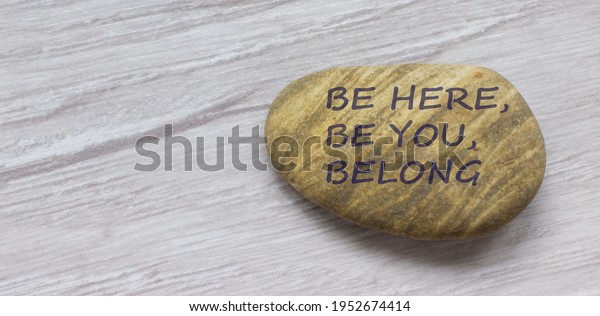 Be here, be\
you, belong symbol. Beautiful stone with words \'Be here, be you,\
belong\' on beautiful white wooden background. Diversity, business,\
inclusion and belonging\
concept.