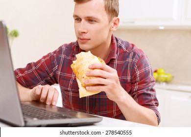 To Be Keen. Handsome Young Man Eating Big Sandwich In Front Of Computer In Kitchen. 