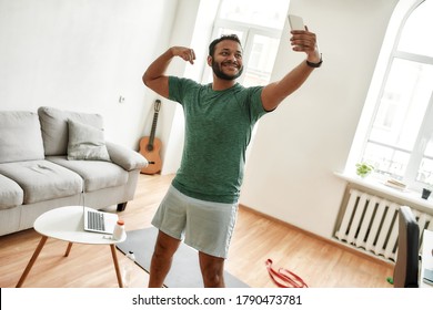 Be Physically Fit. Excited Bearded Active Man Holding Smartphone, Showing Biceps While Taking A Selfie During Morning Workout At Home. Fitness, Motivation Concept. Horizontal Shot