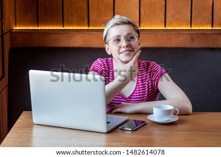 Be waiting an idea! Portrait of thoughtful attractive young girl freelancer with short hair, in pink shirt and eyeglasses is sitting in cafe and working on laptop, try to remember information. indoor
