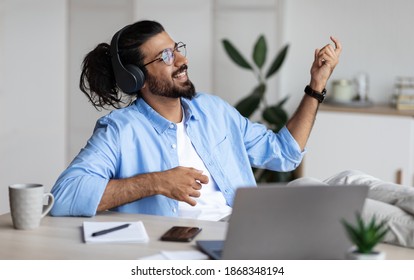 Be Yourself. Joyful weatern freelancer guy listening music in headphones and playing virtual guitar, relaxing at desk in home office, young indian man having fun and enjoying working remotely