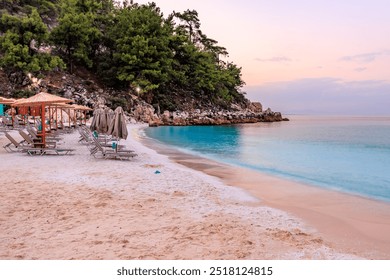 A beach with a beautiful blue ocean and a rocky cliff in the background. There are several umbrellas and chairs set up on the beach for people to relax and enjoy the view. The atmosphere is calm - Powered by Shutterstock