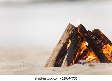 Beach Campfire On Lake With Sand Shore. Burning Wood On White Sand In Daytime 