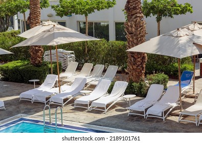 Beach Chairs, Umbrellas Around Open Air Swimming Pool, Metal Handrails, Blue Water With Palm Trees Background. Beach Sun Beds With A White Towels. Empty Lounge Chair In A Row With Umbrella For Relax