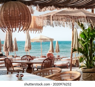 Beach club concept, food corner or cafe, restaurant tables, straw natural chairs, wooden tables, makrome knitted sun umbrellas, white modern sun beds, sea view on background - Shutterstock ID 2020976252