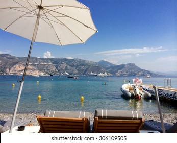 Beach Club In The South Of France/ Paloma Beach/ St Jean Cap Ferrat, France - September 5, 2014: View Of The Mediterranean With Two Empty Lounge Chairs On Paloma Beach With Parasol And Rubber Boat