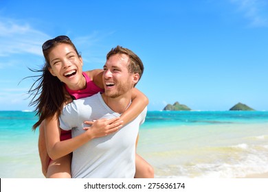 Beach Couple Having Fun Laughing On Hawaii Holiday. Beautiful Asian Mixed Race Woman Piggybacking On Caucasian Boyfriend On Lanikai Beach, Oahu, Hawaii, USA With Mokulua Islands.