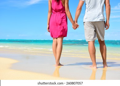 Beach Couple In Love Holding Hands On Honeymoon. Lower Body Crop Showing Pink Dress, Casual Beachwear, Legs And Feet Of Romantic Newlyweds People Standing On White Sand On Travel Summer Vacations.