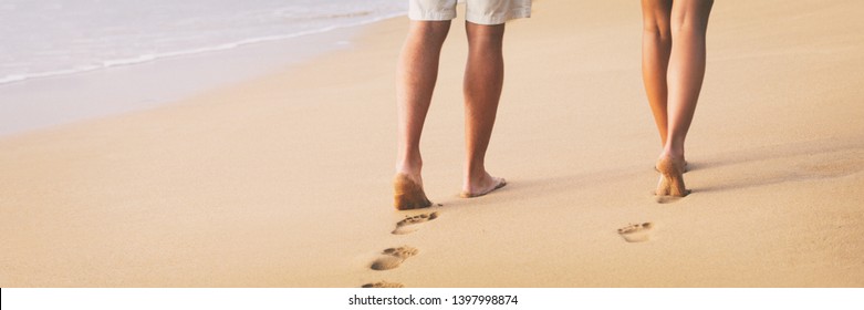 Beach couple walking barefoot on sand at sunset walk honeymoon travel banner - woman and man relaxing together leaving footprints in the sand.