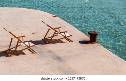 Beach Deck Chairs Facing The Sun At Turquoise Sea And Rusty Bollard At Exclusive Spot