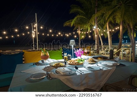 beach dinner table,Table set up for a romantic meal on the beach with lanterns and chairs and flowers with palm trees and sky and sea in the background.