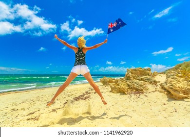 Beach Freedom Summer Vacation. Joyful Happy Woman Jumping On White Beach Waving Australian Flag. Blonde Girl Happiness Jump In Mettams Pool, North Beach Near Perth, Western Australia. Blue Sky.