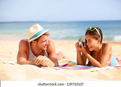 Beach fun couple travel. Woman taking photo picture of man smiling happy with retro vintage camera, Cool trendy modern hipster interracial couple on summer holidays vacation on tropical beach.