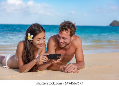 Beach holiday couple sunbathing relaxing having fun using phone app 5g data. Young tourists friends technology lifestyle. Happy multiracial Asian Caucasian biracial family enjoying summer lifestyle. - Powered by Shutterstock