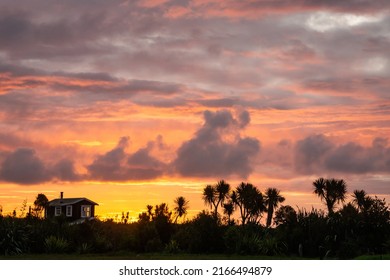 A beach house and cabbage trees are silhouetted on the horizon at dusk with a bright red, orange and yellow skies at Okarito, West Coast, South Island, New Zealand.
