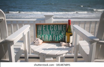 Beach House Porch Overlooking Ocean With Two Chairs In White With A Bottle Of Wine Seashells And Paradise Sign.