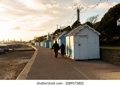 Beach Huts In Felixstowe, Suffolk, England With An Unknown Elderly Couple Walking Away From Camera