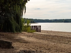 Beach Landscape With Wooden Wave Breakers And Big Trees. The Calm Water Of The Bautzen Reservoir Is Glowing In The Sunlight. The Sandy Bathing Beach In The Front Is Empty. The Sky Is Covered.