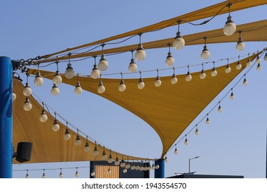 Beach Lighting Equipment. Texture Of Fabric Stretch Roof Made Of Canvas Sail With Rows Of Bulb Lamps For Party Night Lighting On Blue Sky Background.