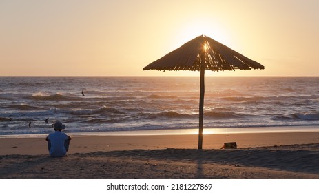 Beach Ocean Waterline Man  Silhouetted Sitting Next To Wood Umbrella Watching Sunrise On The Sea Horizon.