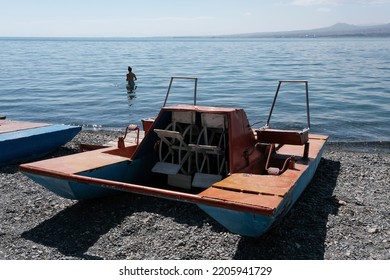Beach on Sevan lake with old paddle boat (pedalo, water bike) on pebble and girl in the water on sunny summer day, Armenia.