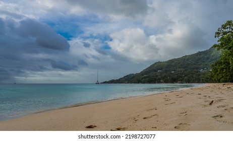 A Beach On A Tropical Island. Footprints In The Sand. The Turquoise Ocean Is Calm. A Green Hill Against A Background Of Blue Sky And Clouds. Seychelles. Mahe. Beau Vallon