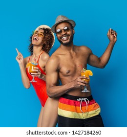 Beach party. Happy black girl and guy in swimwear dancing with summer cocktails, blue studio background - Powered by Shutterstock