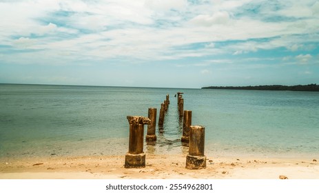 A beach with a pier and a body of water. The pier is old and has a few wooden posts. The water is calm and the sky is blue - Powered by Shutterstock