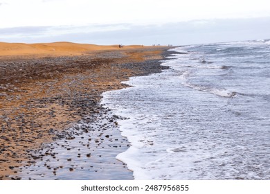 Beach with a rocky shoreline and a few people walking along the beach. The water is calm and the sky is cloudy - Powered by Shutterstock