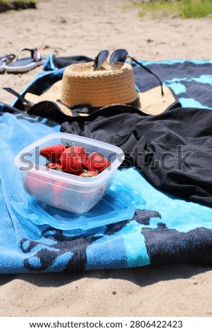 Beach scene featuring a straw hat, sunglasses, black shirt, and a container of strawberries on a blue towel laid out on sandy ground
