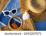 Beach scene with straw, white sunglasses, sunscreen in wicker bag on blue towel. Summer essentials for sun protection on sandy shore by ocean. Relaxing day at beach necessary sunbathing.