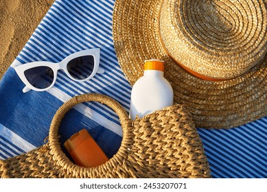 Beach scene with straw, white sunglasses, sunscreen in wicker bag on blue towel. Summer essentials for sun protection on sandy shore by ocean. Relaxing day at beach necessary sunbathing.