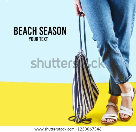 Beach season. Woman with jeans and sandals is holding a beach striped bag on a blue yellow pastel background. Summer time in a sea resort
