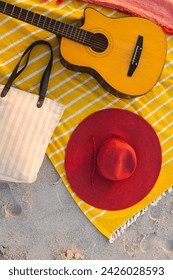 A beach setup features a guitar and a red hat on a yellow blanket. Beach essentials create a relaxing outdoor scene, evoking leisure and musical enjoyment.