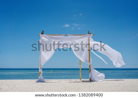 Beach shade with white gracefully fluttering fabric curtains on seashore. Sea breeze and white sand