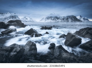 Beach with stones in blurred waves, blue sky with low clouds and snowy mountains at dusk in winter. Sea coast in Lofoten islands, Norway. Dramatic landscape with sea, rocks in fog. Moody scenery - Powered by Shutterstock