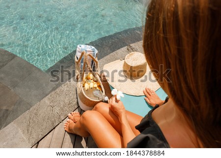 Beach, summer vacation and accessories concept. Woman sit on subned. Her legs near wicker bag with towel, straw hat and still water in glass near the swimming pool.  Summer vacation concept 