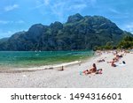 Beach with sunbathing people at lake in mountains, cliffs and blue sky in background, Lago di Garda, Riva del Garda, Italy