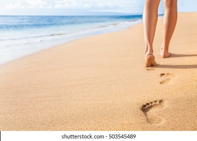 Beach travel - woman relaxing walking on sand beach leaving footprints in the sand. Closeup detail of female feet and legs on golden sand on Kaanapali beach, Maui, Hawaii, USA.