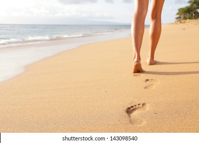 Beach travel - woman walking on sand beach leaving footprints in the sand. Closeup detail of female feet and golden sand on Kaanapali beach, Maui, Hawaii, USA.