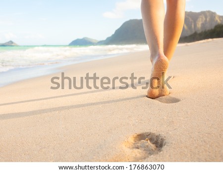 Beach travel - woman walking on sand beach leaving footprints in the sand. Closeup detail of female feet and golden sand on beach in Hawaii.